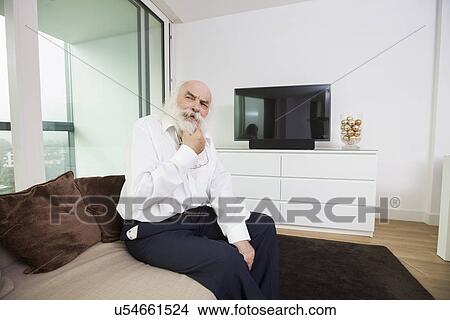 Thoughtful senior man sitting on sofa in living room View Large Photo Image Picture - Thoughtful senior man sitting on sofa in living room. Fotosearch