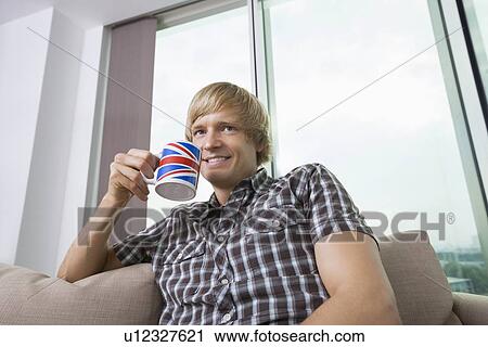 Stock Image - Thoughtful smiling mid-adult man with coffee cup in living room at home. Fotosearch