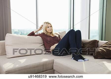 Thoughtful young woman sitting on sofa against window at home View Large Photo Image Stock Photo - Thoughtful young woman sitting on sofa against window at home. Fotosearch