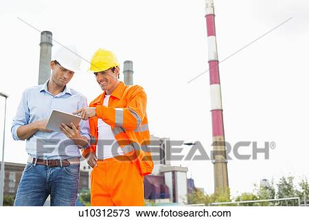 Stock Image - Two construction workers discussing over tablet PC at industry. Fotosearch