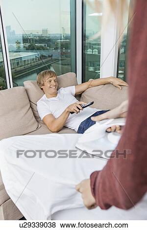 Woman ironing shirt while happy man watching TV on sofa at home View Large Photo Image Stock Photo - Woman ironing shirt while happy man watching TV on sofa at home. Fotosearch