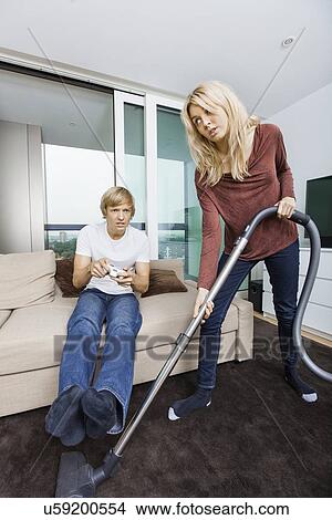 Picture - Woman vacuuming while man play video game in living room at home. Fotosearch