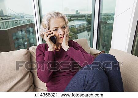Stock Image - Worried senior woman answering smart phone on sofa at home. Fotosearch