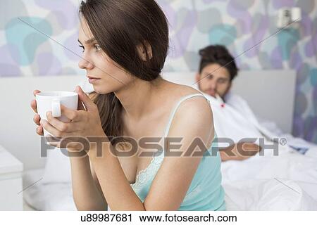 Stock Image - Worried young woman holding coffee mug while ill man relaxing in background. Fotosearch