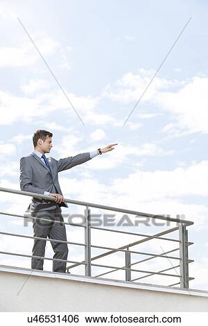 Young businessman with arm raised standing at terrace railings against sky View Large Photo Image Stock Photograph - Young businessman with arm raised standing at terrace railings against sky. Fotosearch