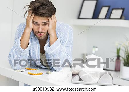 Stock Image - Young ill man suffering from headache at kitchen counter. Fotosearch