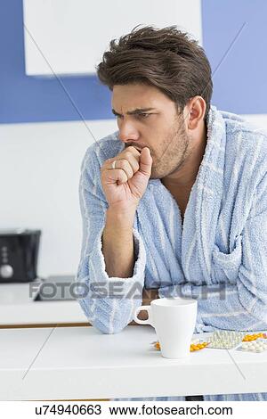 Stock Image - Young man coughing with coffee mug and medicine on kitchen counter. Fotosearch