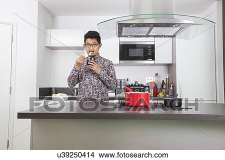Picture - Young man eating broccoli at kitchen counter in home. Fotosearch