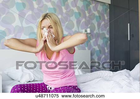 Stock Photography - Young woman blowing her nose in tissue paper while sitting on bed. Fotosearch
