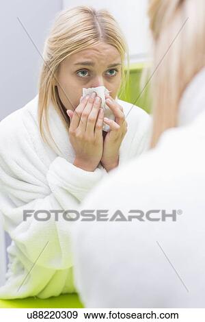 Stock Photo - Young woman blowing nose in tissue paper while looking at mirror. Fotosearch