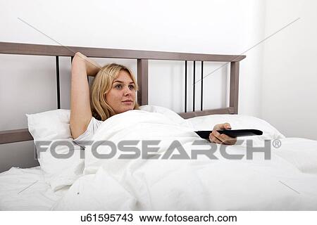 Stock Image - Young woman changing channels with remote control in bed. Fotosearch