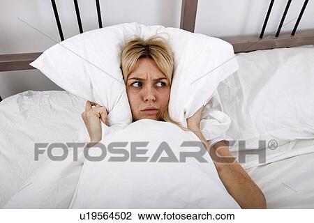 Stock Image - Young woman covering ears with pillow in bed. Fotosearch