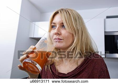 Stock Image - Young woman drinking carrot juice in kitchen. Fotosearch