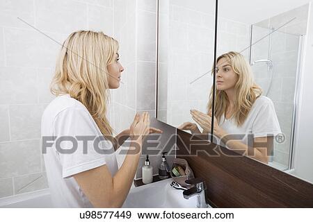 Stock Photo - Young woman looking at mirror in bathroom. Fotosearch