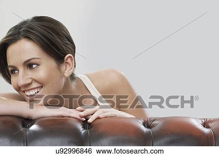 Stock Photograph - Young woman looking away while leaning on sofa against gray background. Fotosearch