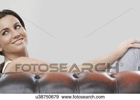 Stock Photo - Young woman sitting on sofa against gray background. Fotosearch