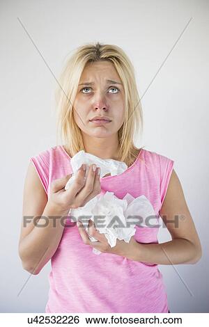 Stock Photograph - Young woman suffering from cold looking up while holding tissues against gray background. Fotosearch