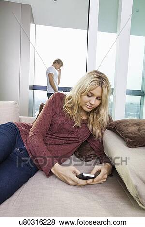 Young woman text messaging while man using cell phone in background at home View Large Photo Image Stock Photo - Young woman text messaging while man using cell phone in background at home. Fotosearch