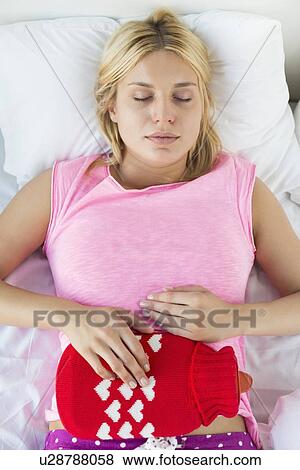 Stock Photo - Young woman using hot water bottle on abdomen while lying on bed. Fotosearch