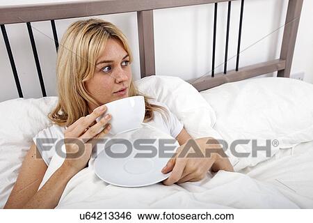 Stock Photograph - Young woman with coffee cup looking away in bed. Fotosearch