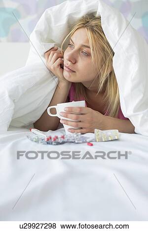 Stock Photo - Young woman with coffee mug and medicines suffering from fever while covered in quilt on bed. Fotosearch