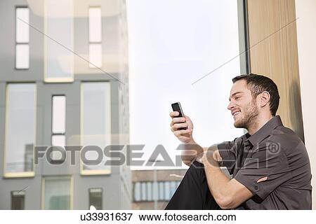 Businessman sitting on windowsill using smartphone View Large Photo Image Stock Photo - Businessman sitting on windowsill using smartphone. Fotosearch