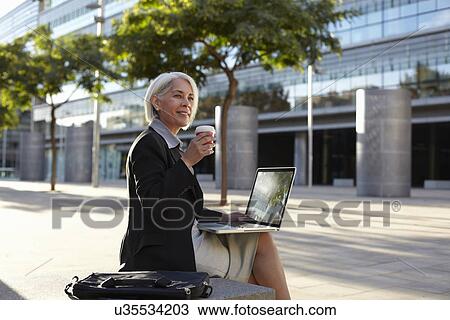Stock Image - Businesswoman sitting outside using laptop and drinking hot drink. Fotosearch