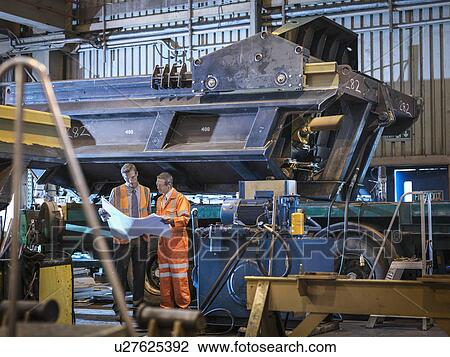 Stock Image - Engineers inspecting engineering drawings together in factory. Fotosearch