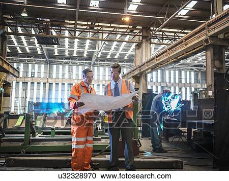 Stock Image - Engineers inspecting engineering drawings in factory. Fotosearch