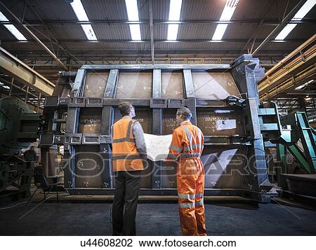 Stock Image - Engineers inspecting engineering drawings in factory, rear view. Fotosearch