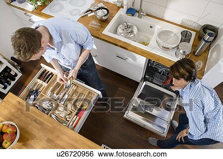 Stock Photograph - Grandmother and adult grandson tidying away cutlery from dishwasher. Fotosearch