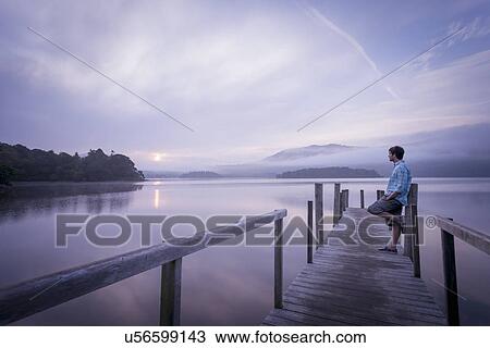 Stock Image - Man on pier by tranquil lake, Cumbria, England, UK. Fotosearch