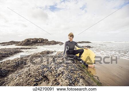 Stock Image - Mature female bodyboarder sitting on rocks, Devon, UK. Fotosearch