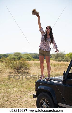 Stock Photo - Mid adult women standing on hood of car waving hat. Fotosearch