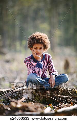 Stock Image - Portrait of boy sitting cross legged on tree stump. Fotosearch