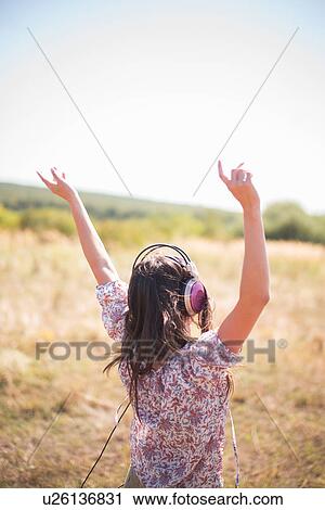 Stock Image - Portrait of mid adult woman dancing in field with arms raised, wearing headphones. Fotosearch