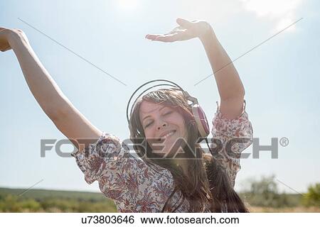 Stock Photograph - Portrait of mid adult woman dancing in field wearing headphones with arms raised. Fotosearch