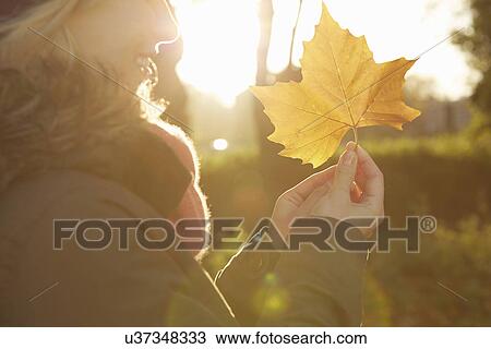 Stock Image - Portrait of young woman in park, holding autumn leaf. Fotosearch