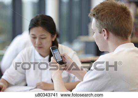 Stock Image - Teenage boy using cell phone in classroom. Fotosearch