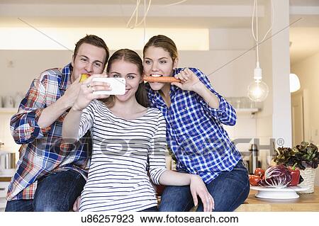 Stock Image - Three young people having fun taking photographs in the kitchen. Fotosearch