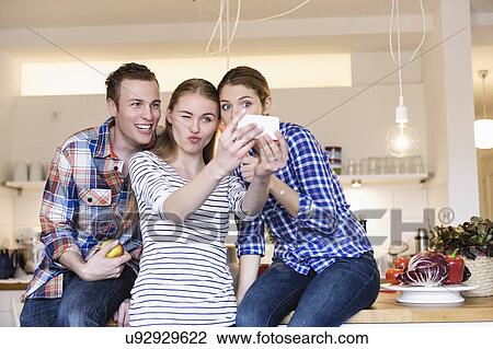 Stock Image - Three young people having fun taking photographs in the kitchen. Fotosearch