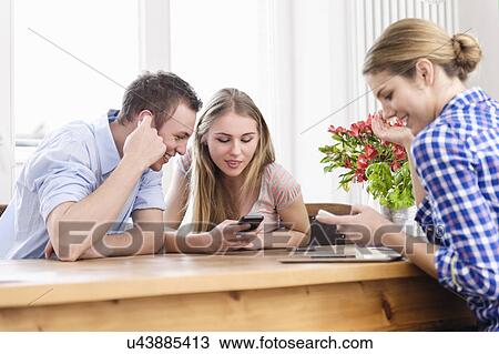 Stock Image - Three young people sitting at a table using cell phones and smiling. Fotosearch