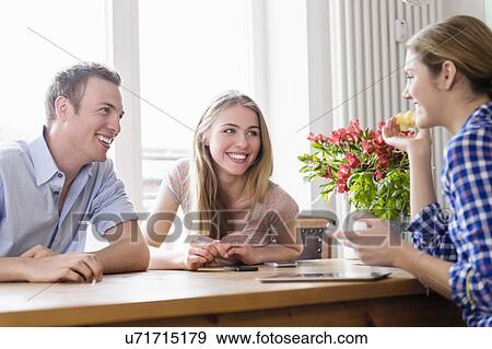 Stock Photo - Three young people sitting at a table talking and smiling. Fotosearch