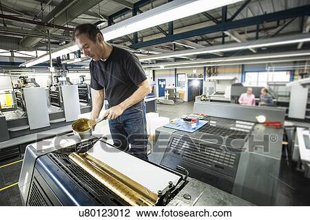 Stock Image - Worker applying gold ink to printing machine in print workshop. Fotosearch
