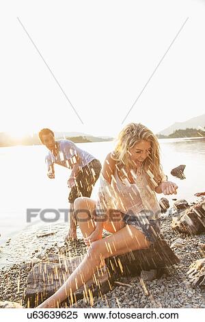 Stock Photography - Young couple by lake, man splashing woman, Cumbria, England, UK. Fotosearch