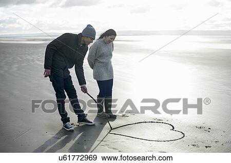 Stock Photo - Young couple drawing heart in sand, Brean Sands, Somerset, England. Fotosearch
