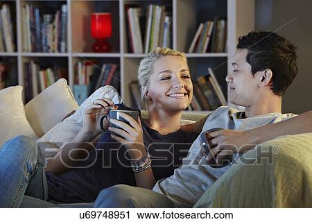 Stock Image - Young couple sitting on sofa with hot drinks. Fotosearch