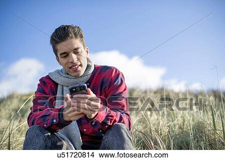 Young man sitting on grass using smartphone View Large Photo Image Picture - Young man sitting on grass using smartphone. Fotosearch