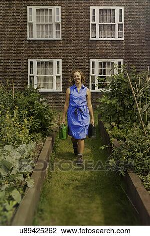 Stock Image - Young woman carrying watering can on council estate allotment. Fotosearch