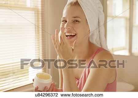 Stock Image - Young woman in bathroom putting on face cream. Fotosearch